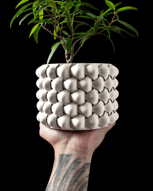Modern 3D printed planter pot in matte white, featuring a tightly interwoven surface resembling braided fabric; the thick structure offers a sculptural cylindrical form with a bold, repetitive pattern of diamond-shaped interlocks, seen held in a tattooed hand against a black background, showcasing its size and texture with a living plant inside.