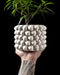 Modern 3D printed planter pot in matte white, featuring a tightly interwoven surface resembling braided fabric; the thick structure offers a sculptural cylindrical form with a bold, repetitive pattern of diamond-shaped interlocks, seen held in a tattooed hand against a black background, showcasing its size and texture with a living plant inside.