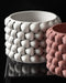 Two cylindrical 3D printed planters with a bold braided texture; the white and terracotta variants are displayed on a glossy black surface, with the white one in focus showing deeply carved interwoven geometry and precise, smooth rim.