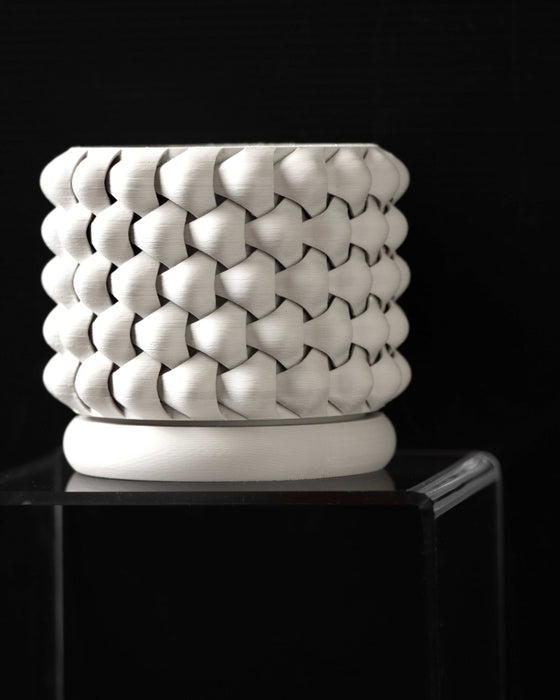 White 3D printed planter placed on a matching base, showcasing the lower curvature of the pot and deep interlocking pattern, with a softly diffused light accentuating the woven texture and sharp definition of each layered segment.