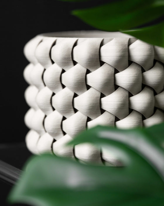 Side view of 3D printed white planter partially hidden behind green leaves; dramatic lighting highlights the three-dimensional braided structure of the pot, creating a play of light and shadow over the raised interlocked texture.