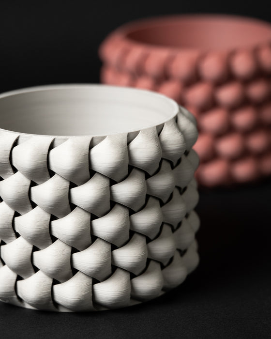 White and terracotta 3D printed planters with matching interwoven texture displayed side by side; the focus is on the foreground white planter showing deep, sculpted layers and a smooth upper rim.