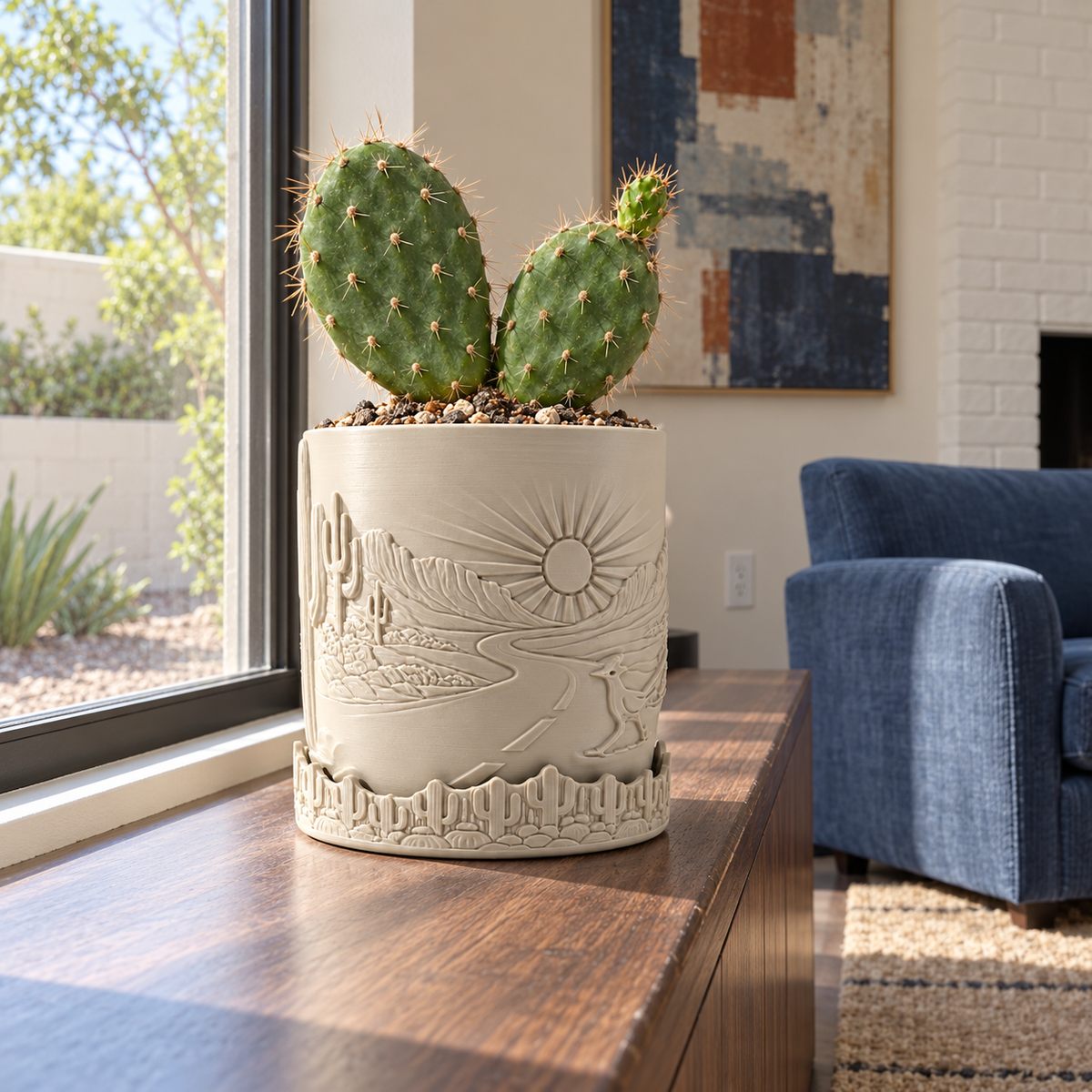 Cactus in a cream Desert Sunrise relief planter on a wooden table beside a window and blue chair.