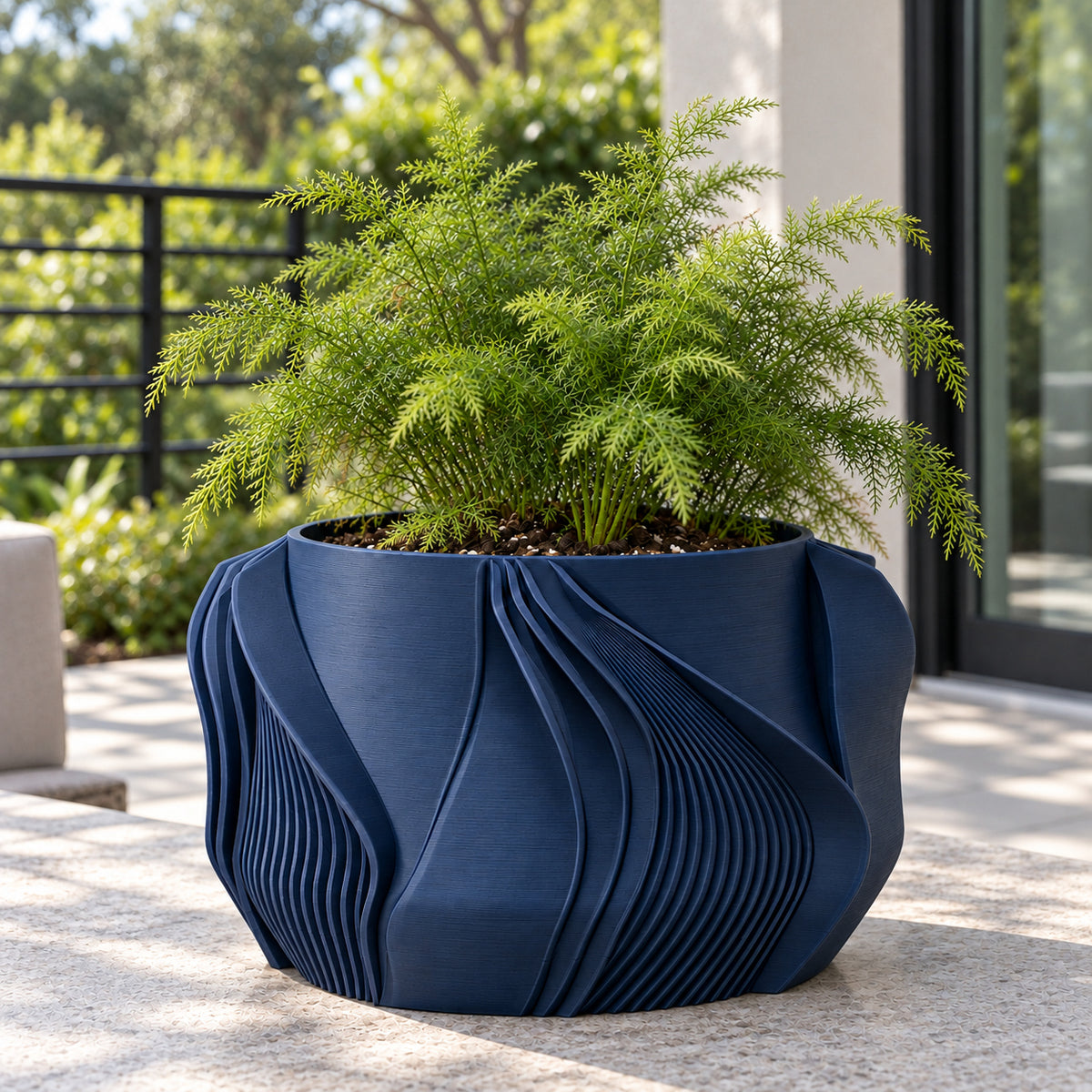 Navy blue Fano planter with green foliage on a sunlit patio beside a railing.