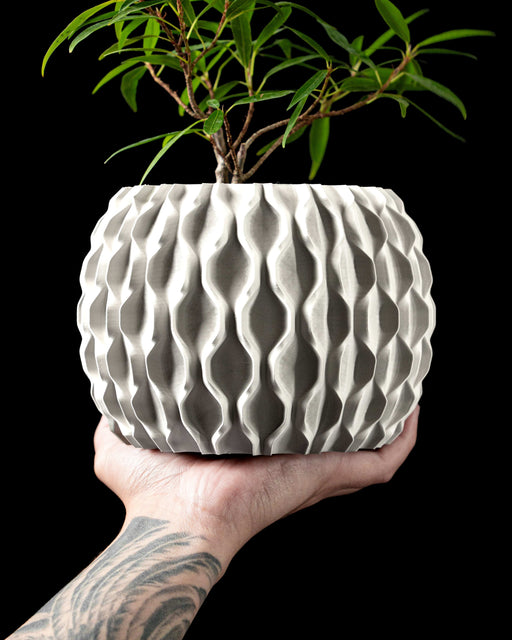 3D printed planter with a cylindrical body and tactile matte surface texture, designed in a muted gray tone, featuring a seamless top section with lightly ribbed vertical grain that mimics handcrafted stone. The base of the pot is elevated on a hollow lattice structure resembling cellular geometry, adding a sculptural feel. The symmetrical contrast between the solid top and openwork base creates balance in contemporary interior styling.