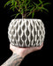 3D printed planter with a cylindrical body and tactile matte surface texture, designed in a muted gray tone, featuring a seamless top section with lightly ribbed vertical grain that mimics handcrafted stone. The base of the pot is elevated on a hollow lattice structure resembling cellular geometry, adding a sculptural feel. The symmetrical contrast between the solid top and openwork base creates balance in contemporary interior styling.