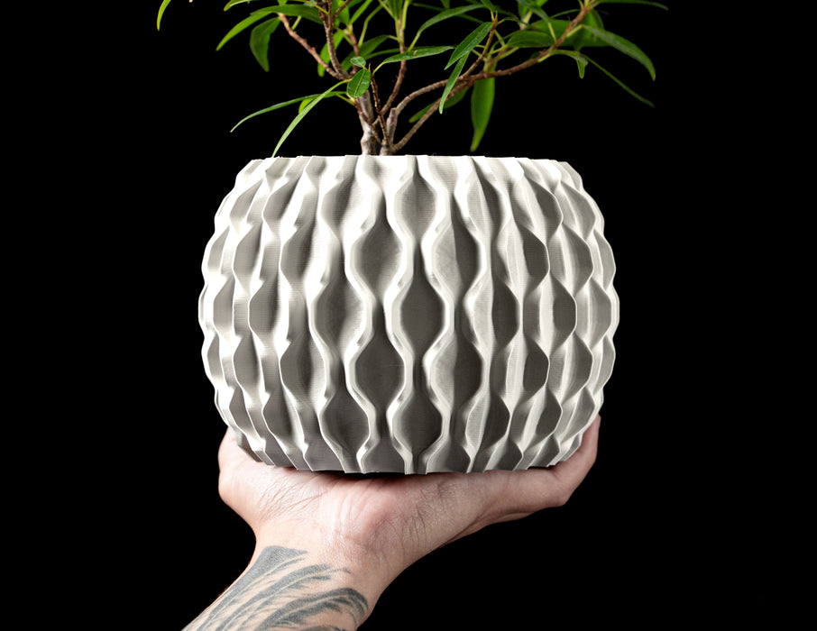 Close-up of a gray cylindrical planter showcasing the detailed texture on the upper container's surface, reminiscent of brushed ceramic but achieved through high-precision 3D printing. The pot is supported by a ring-shaped geometric base with polygonal voids, engineered for both structural elevation and aesthetic intrigue. The minimalist body meets a mathematically inspired foundation, blending organic and digital design principles in a compact form.