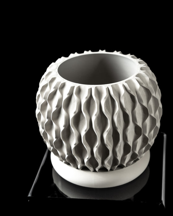 High-contrast image of a matte gray planter viewed from above, revealing the depth of its cylindrical container and beveled rim. Its upper section showcases a fine-grained texture that mimics natural stone, while the lower platform incorporates polygonal architectural openings. The planter exemplifies how 3D printing allows form and function to merge in a single sculptural object ideal for modern and industrial-inspired settings.