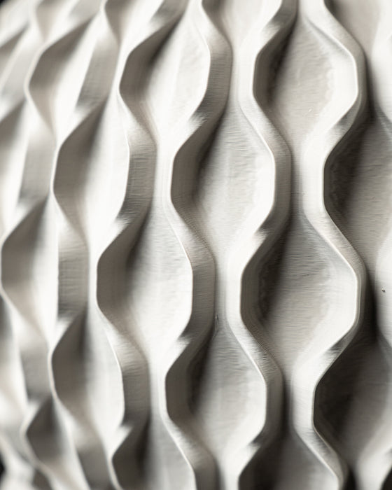 The planter is displayed in a tattooed hand, showcasing its size and scale relative to human touch. The top portion features a vertically brushed gray finish, subtly catching light, while the base remains boldly geometric, evoking a sense of balance between digital form and artisanal tactility. The solid matte finish complements the voided lattice, making this planter an expressive and functional decor item for any surface.