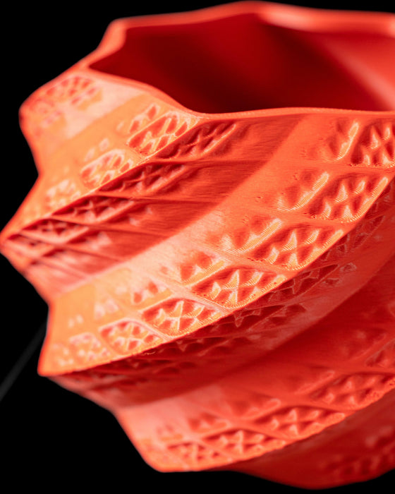 A striking coral-colored 3D printed planter pot sits on a black platform, holding a small bonsai. This ornate flower pot features a spiral lattice design, making it perfect for indoor planters or modern home decor. Ideal as a book plant pot or a statement piece among other flower pots, it’s a unique planter for those who love contemporary style and thoughtful gifts for mom.