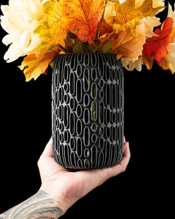 3d printed black vase with intricate vertical oval cutouts arranged in an organic pattern, modern flower vase with futuristic relief design, cylindrical silhouette and rounded base, perfect for dried flowers or decorative stems, unique home decor piece for table or shelf, matte black color highlighting sculptural contours, contemporary vase with detailed textures and structural harmony, vase dried flowers, home office decor, ikebana vase, black vase, vases for flowers, modern vase