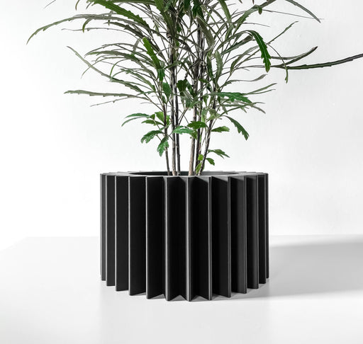 Black 3D printed planter pot with sculptural vertical ridges, cylindrical shape, and a thick top rim – indoor decorative container shown with plant inside
